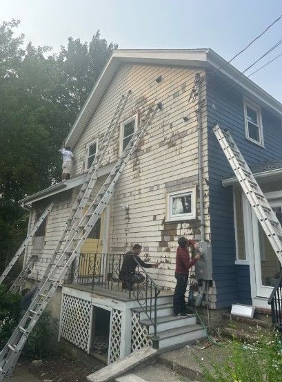 People painting a two-story house, using multiple ladders to reach different sections of the siding; old paint is peeling.