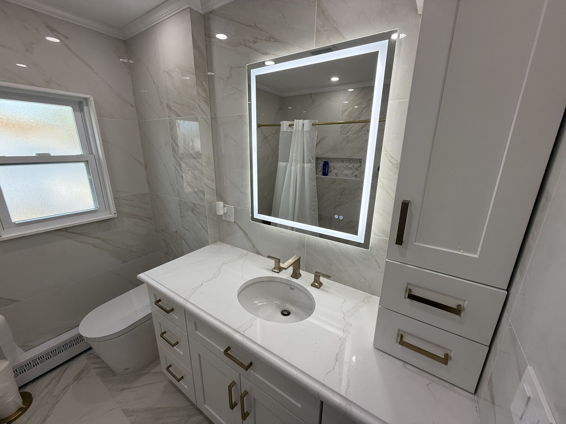 Modern white bathroom with vanity, lit mirror, sink, toilet, and frosted window
