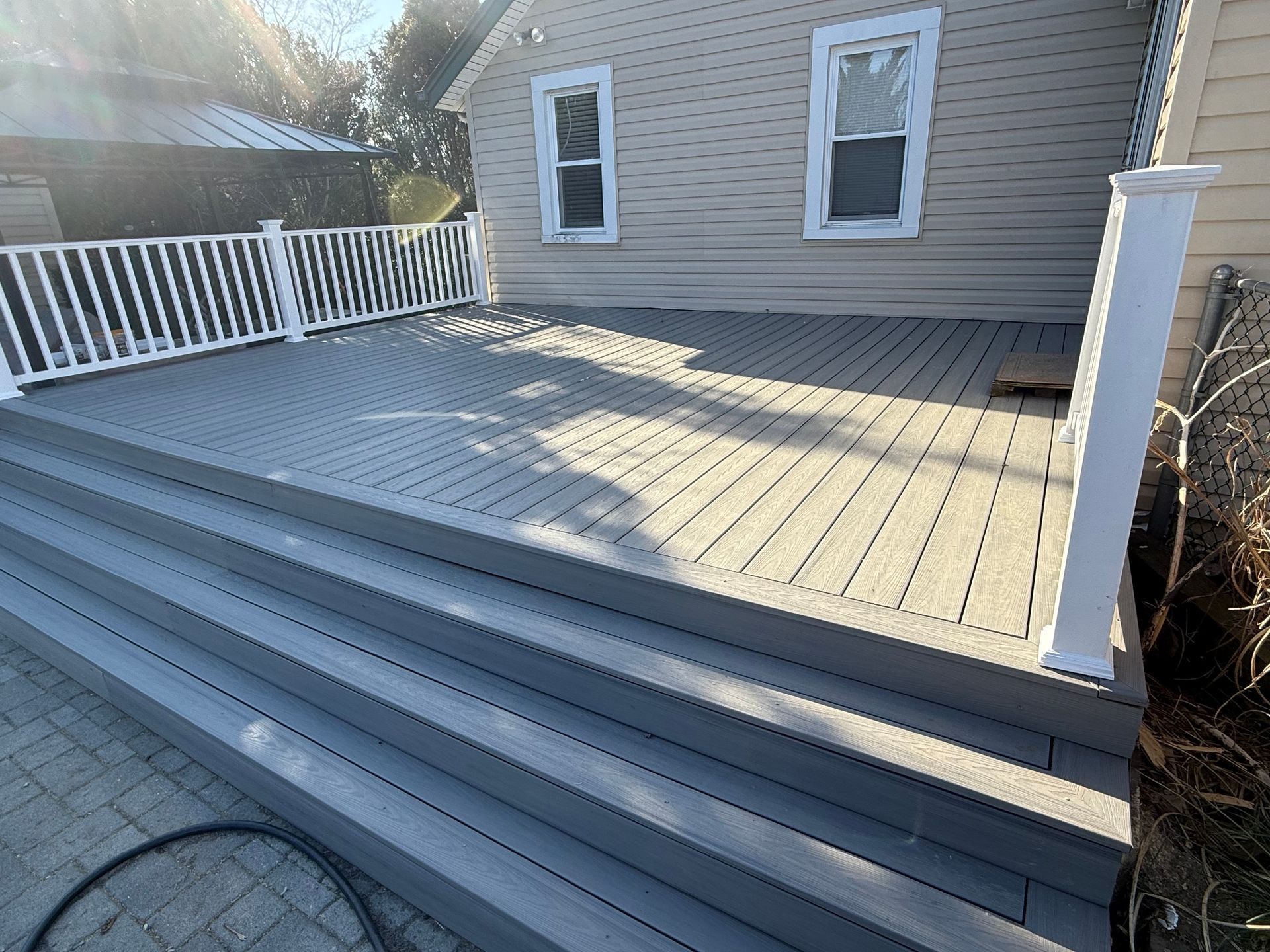 Gray wooden deck with white railings and steps beside a house, lit by sunlight