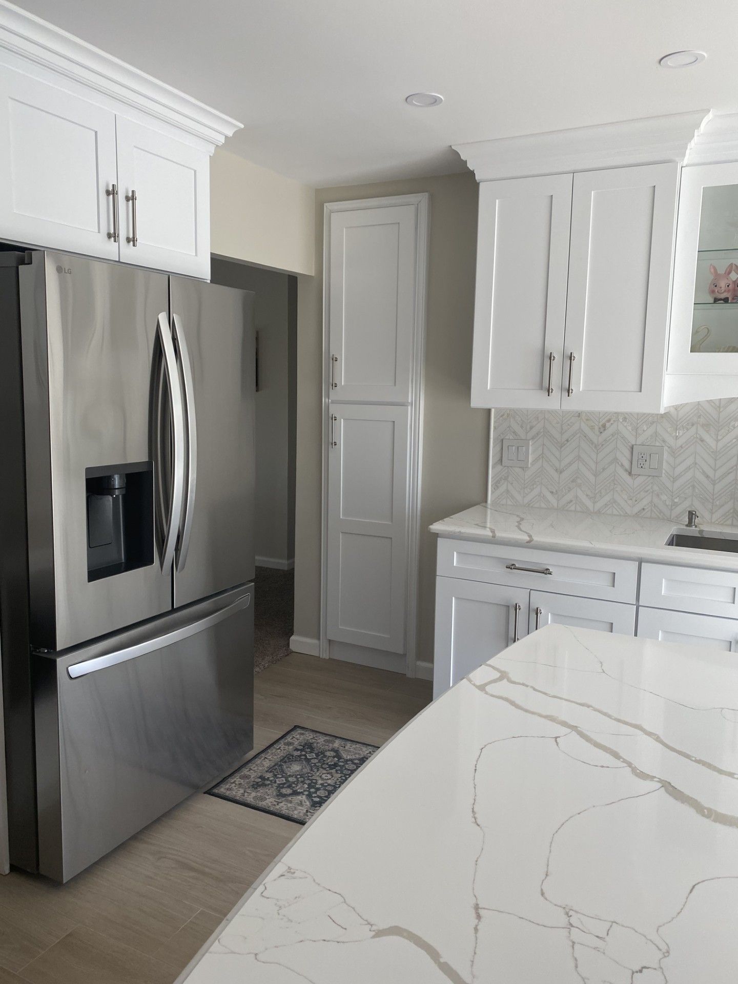 A modern kitchen featuring a stainless steel refrigerator, white cabinets, a narrow pantry, and a marble-patterned island.