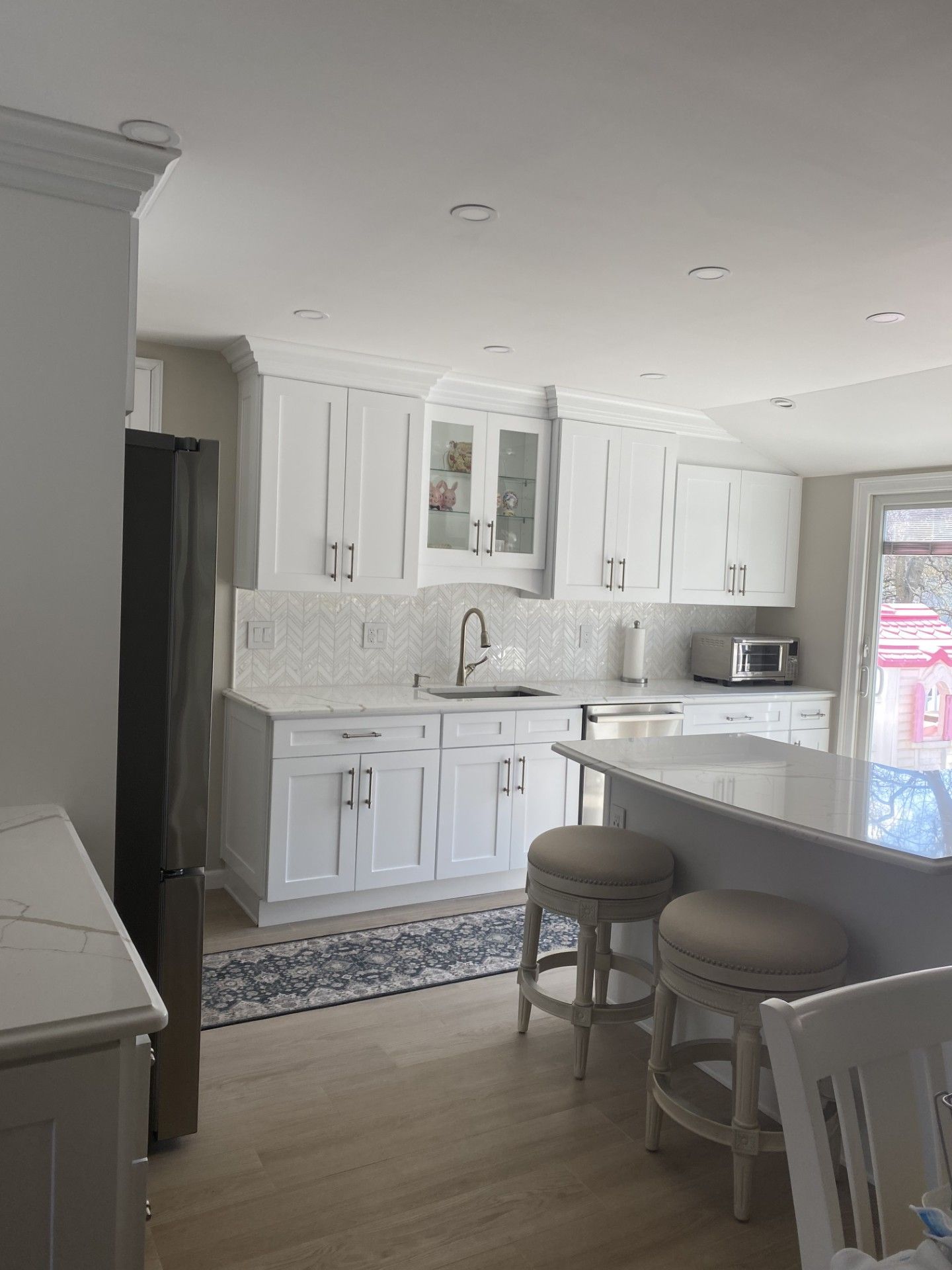 A bright kitchen featuring white cabinets, a light-toned island with two stools, light wood floors, and a patterned rug.