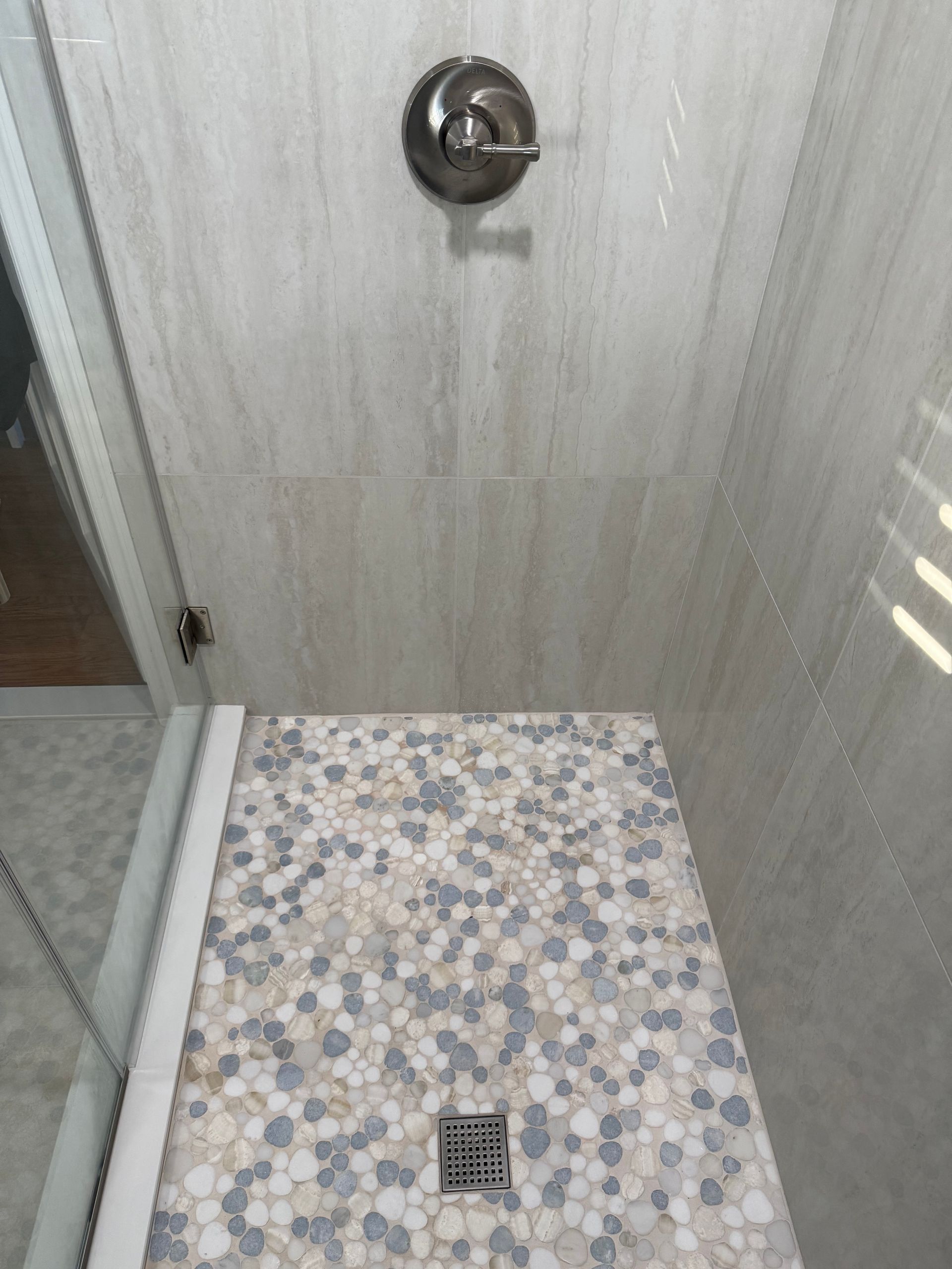 Shower with pebble tile floor and light gray wall tile, brushed nickel fixtures, and glass door.