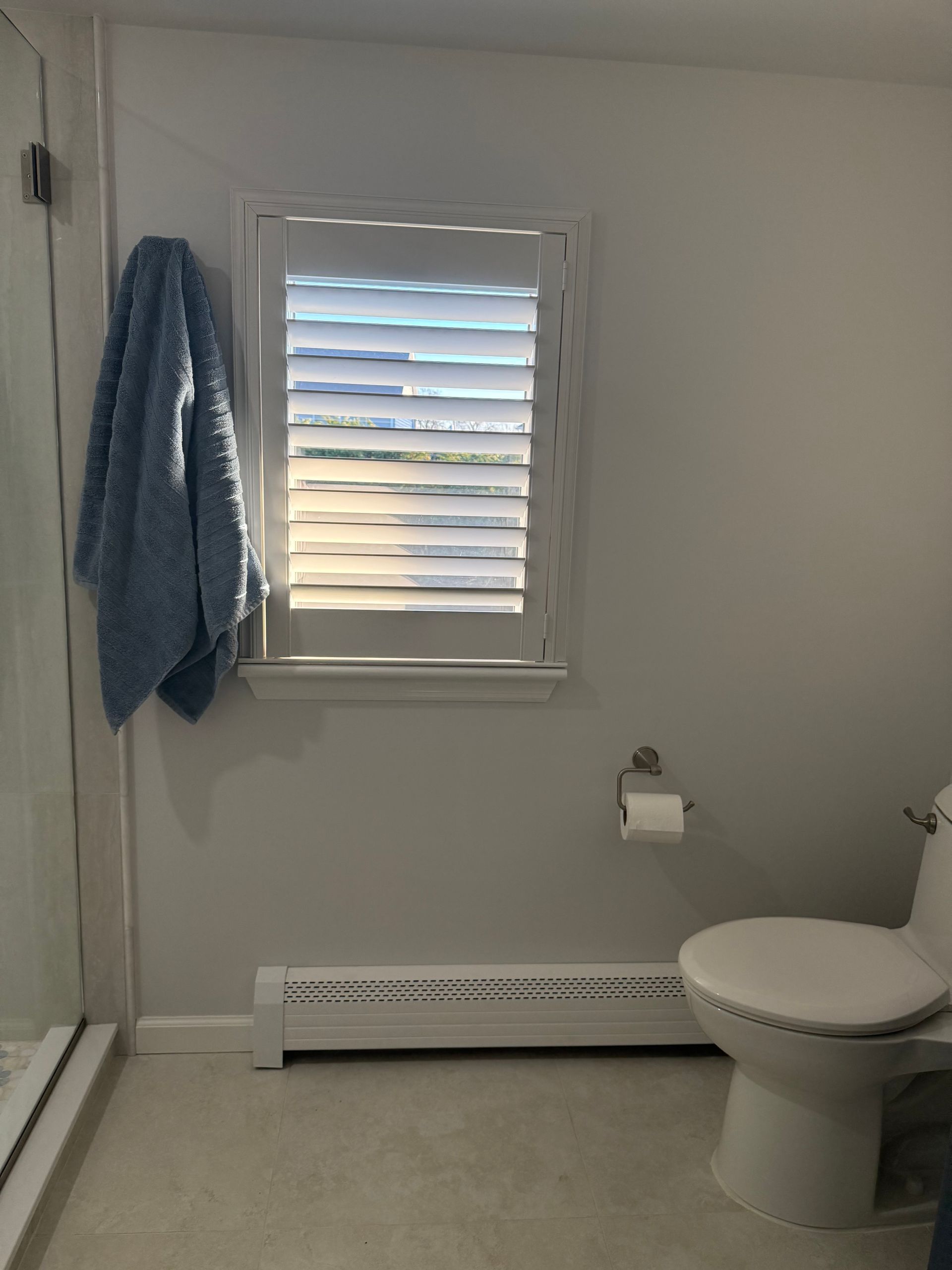 Bathroom with a window, toilet, and a blue towel. White walls and shutters.