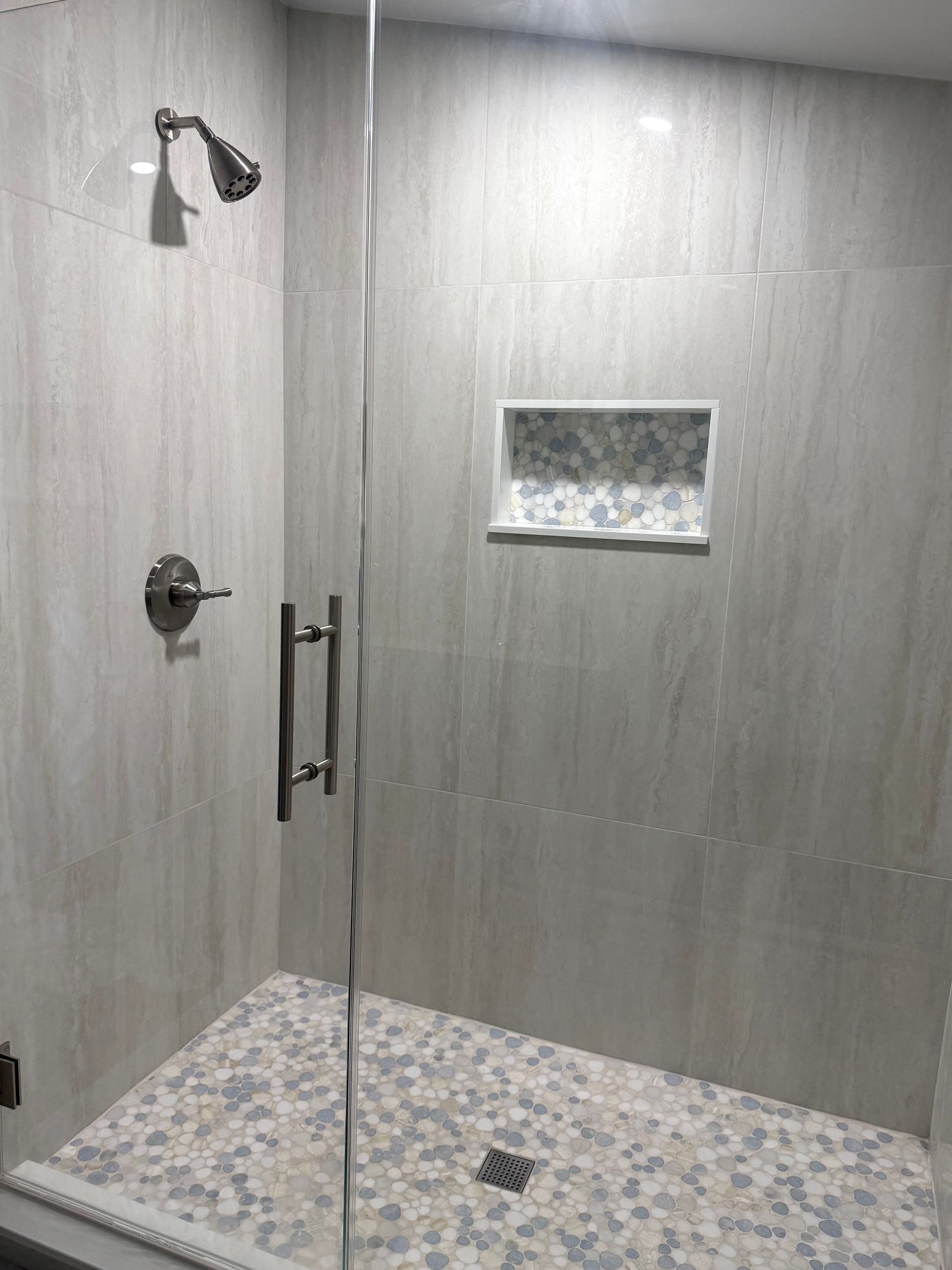 Shower with pebble tile floor, gray wall tiles, glass door, and built-in shelf.