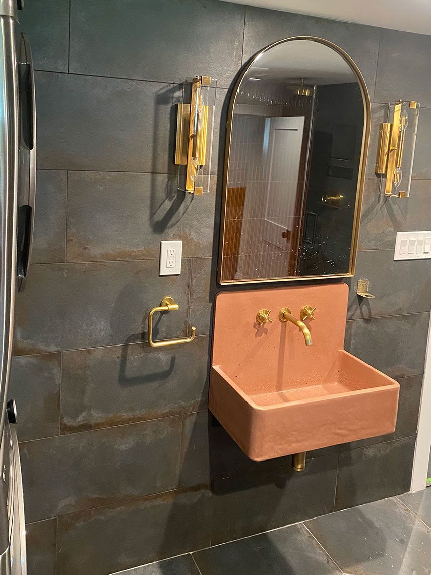A bathroom with a pink sink, gold fixtures, arched mirror, and dark tile walls.
