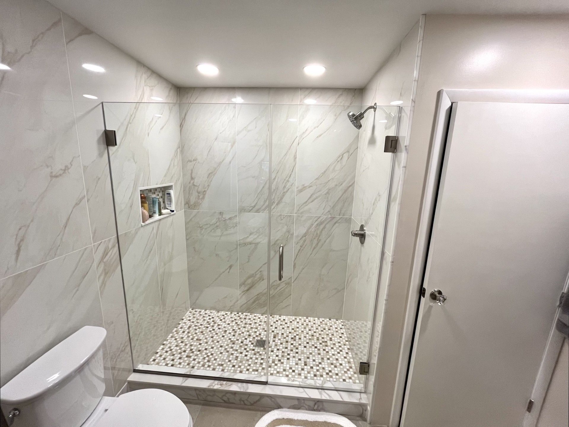 Bathroom with a glass shower, toilet, and marble-look tiles. The shower floor is covered in small stones.