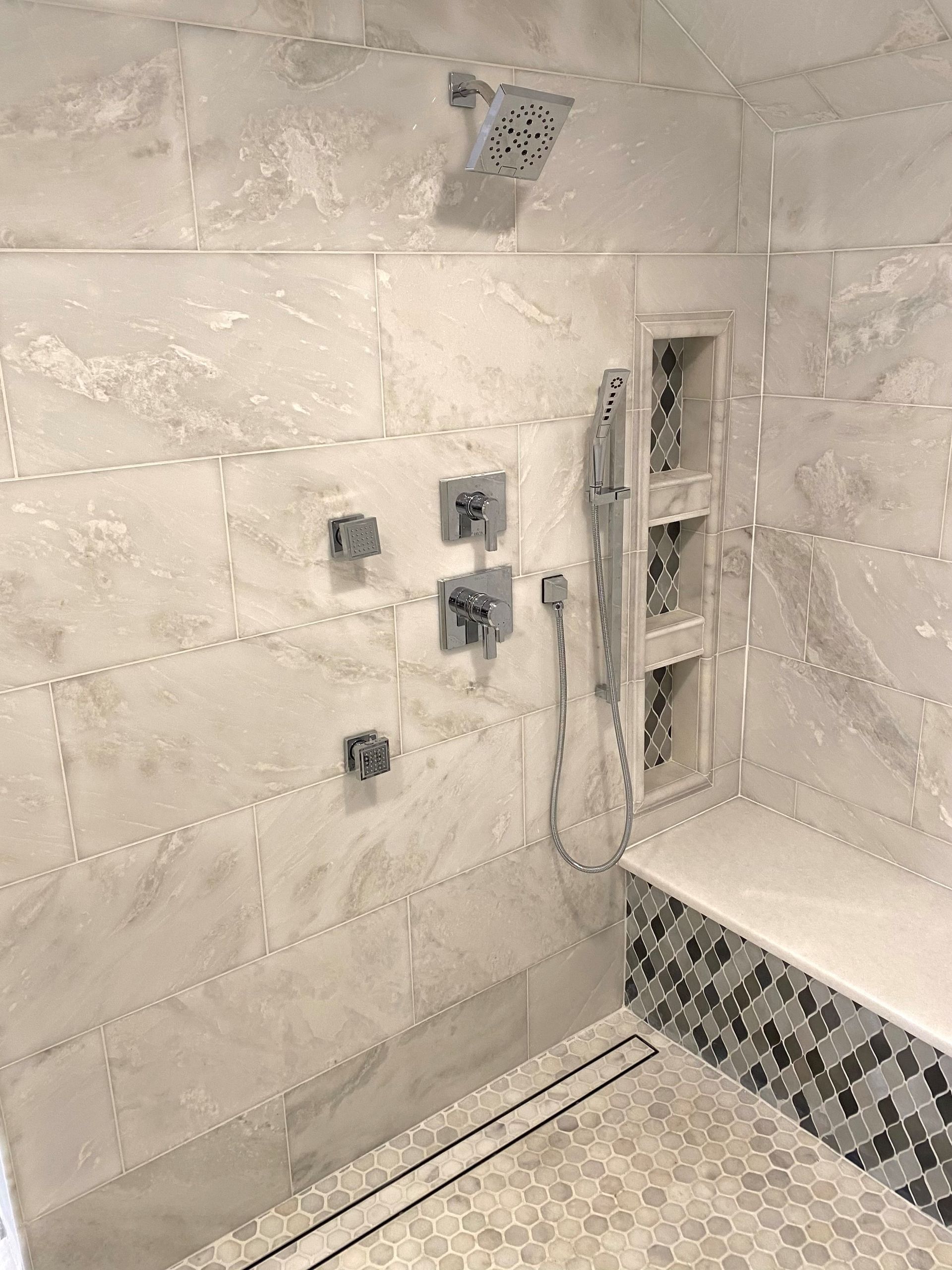 Shower with gray and white tile, built-in bench with mosaic tile accent, and multiple shower heads and controls.