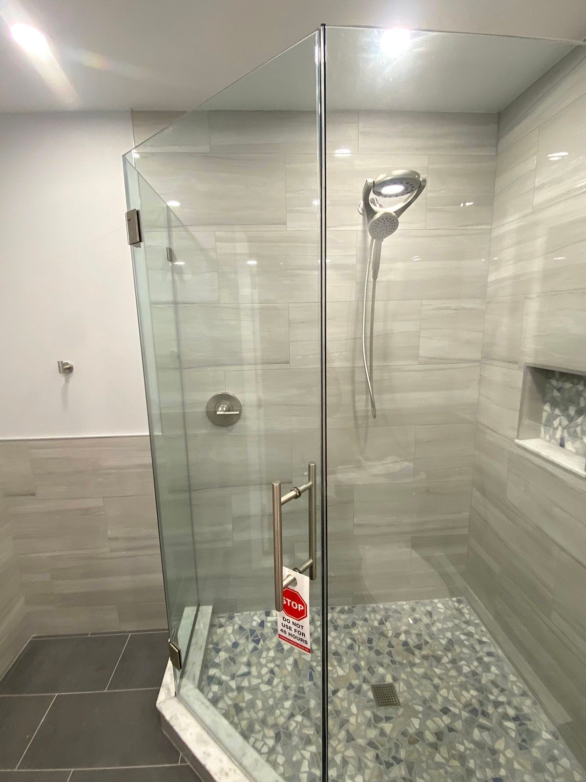 Glass-enclosed shower with gray tiled walls and floor, pebble shower base, and silver fixtures.