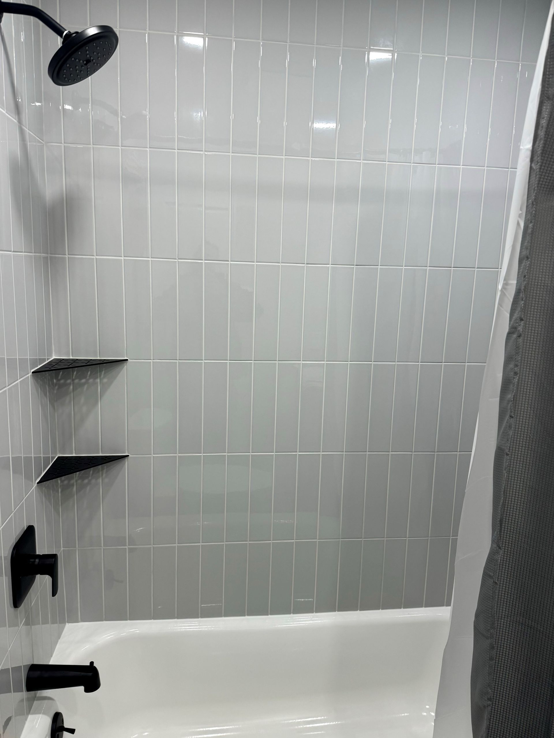 Gray tiled shower with black fixtures and built-in shelves above a white bathtub.