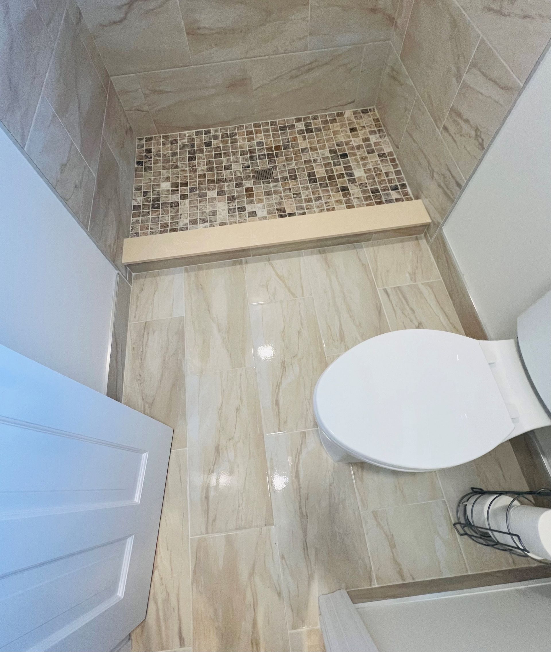 Bathroom with beige tiled floor and walls. Toilet on right, shower with mosaic tile.
