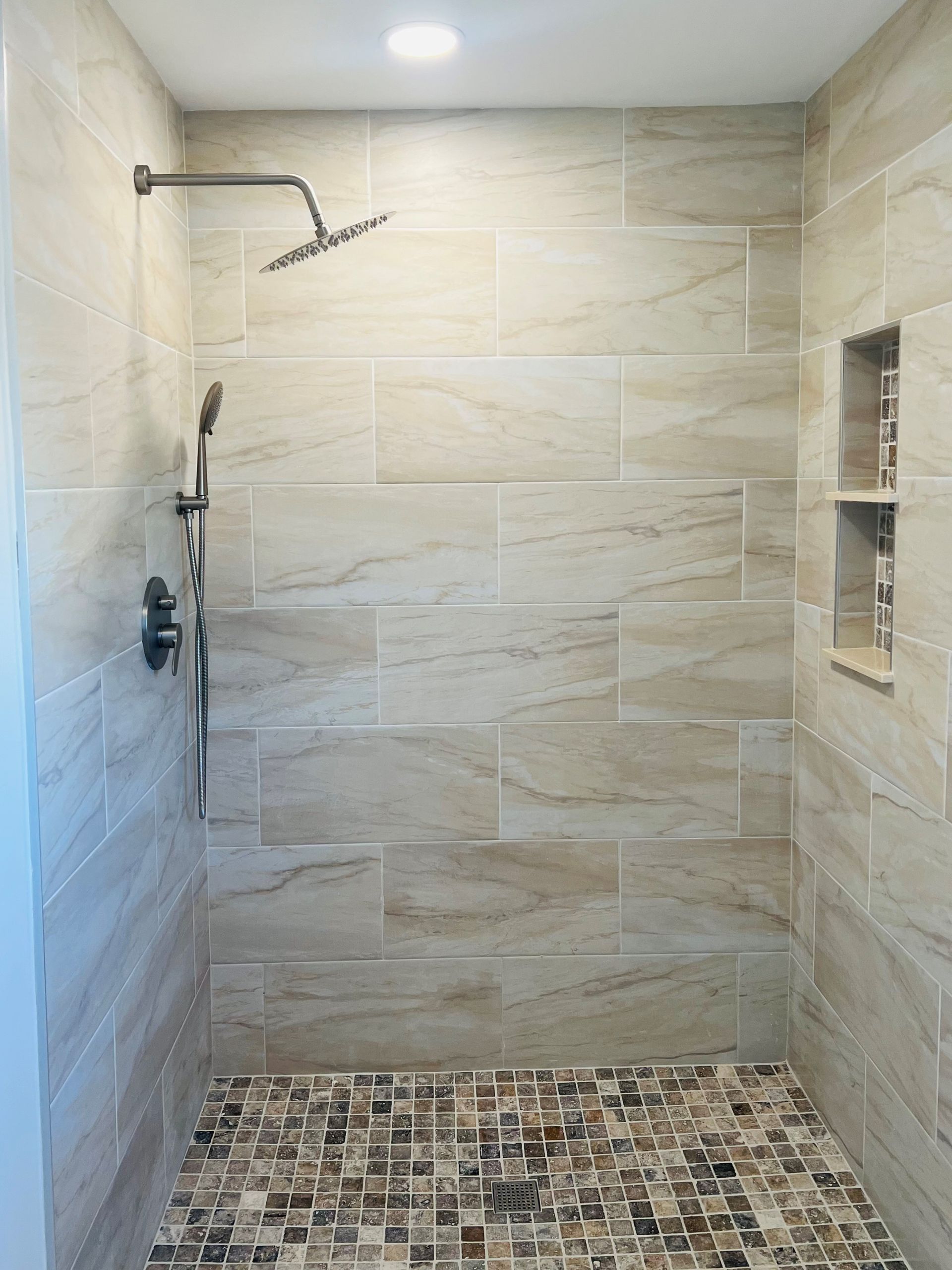 Shower with beige tiled walls, dark shower fixtures, mosaic floor, and recessed shelf.