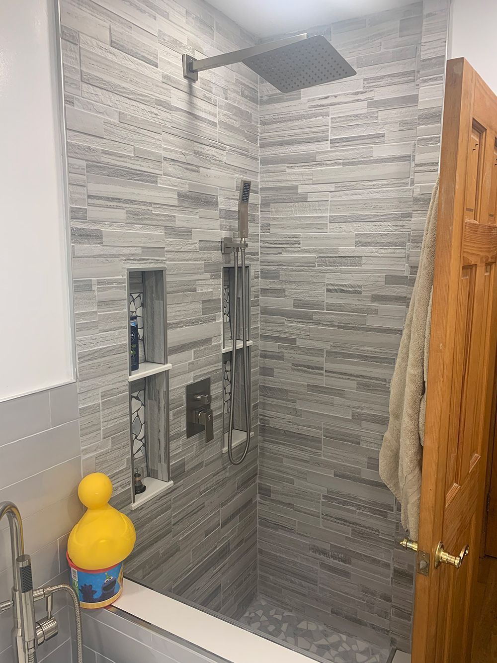 Shower stall with gray tile, recessed shelves, and rain shower head; a wooden door on the right.