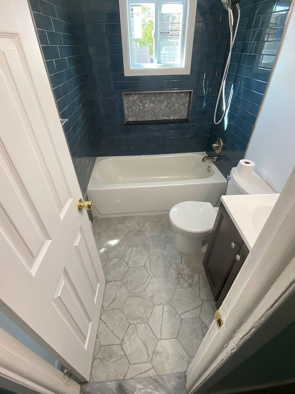 Bathroom with blue tiled shower, white tub and toilet, gray geometric floor tiles.