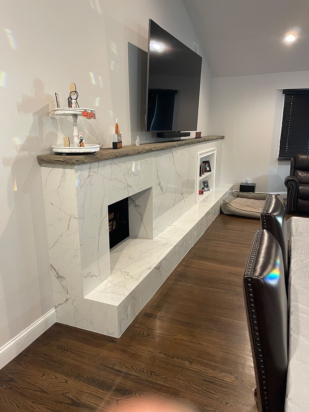 Fireplace with marble-like facade, built-in shelf for TV, figurines, and storage. Brown hardwood floor.