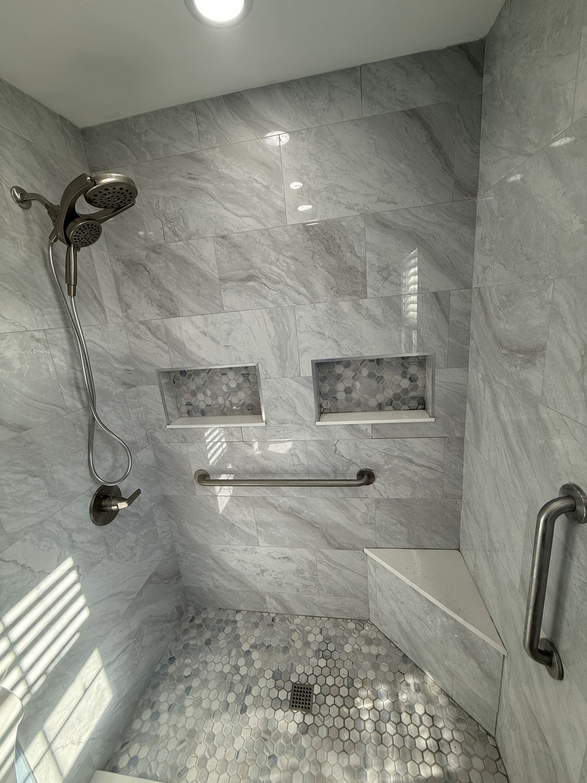 Gray tiled shower with handheld showerhead, grab bars, and built-in shelves.
