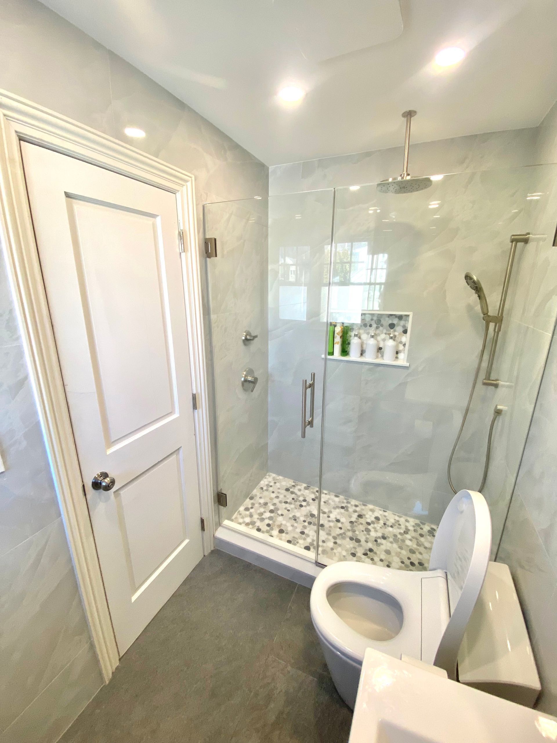 Bathroom with a shower, toilet, and a closed white door. Light grey tiled walls and floor.