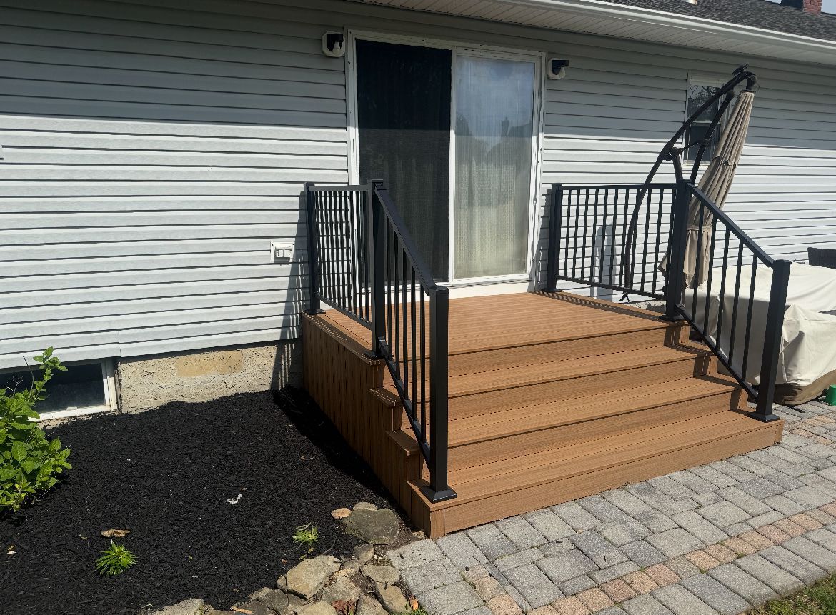 Composite deck stairs with black railing leading to a sliding door of a house with grey siding.