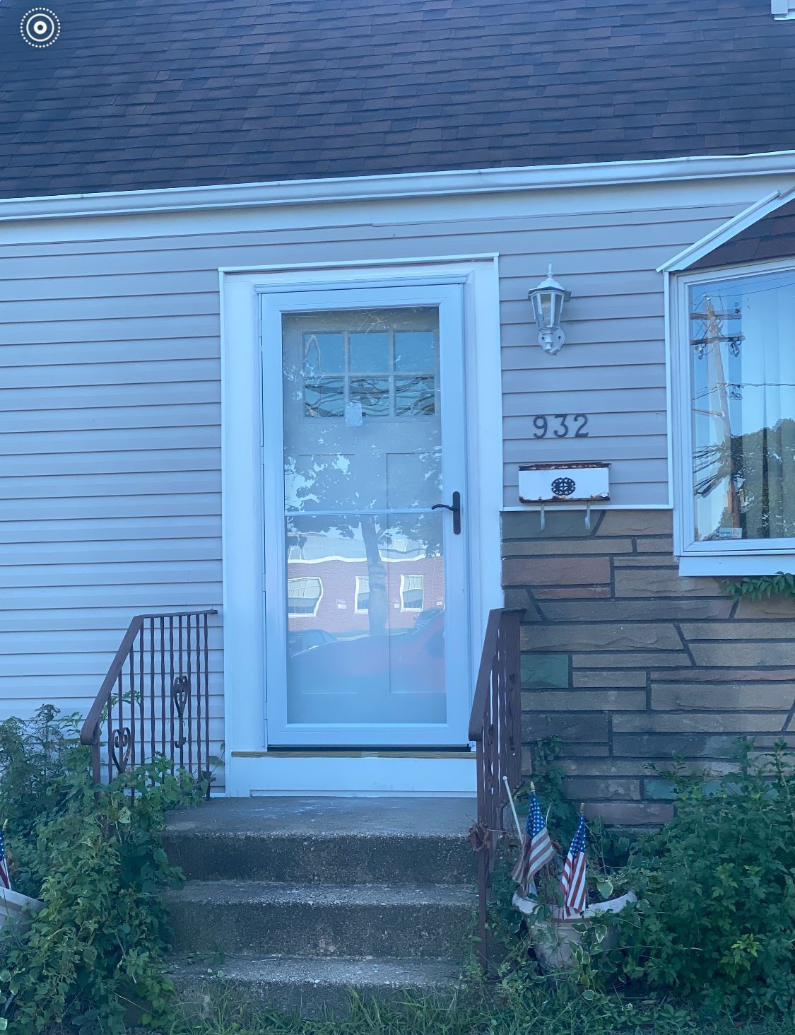 Front door of a house with the number 932. Light blue siding, porch with steps, and bushes.