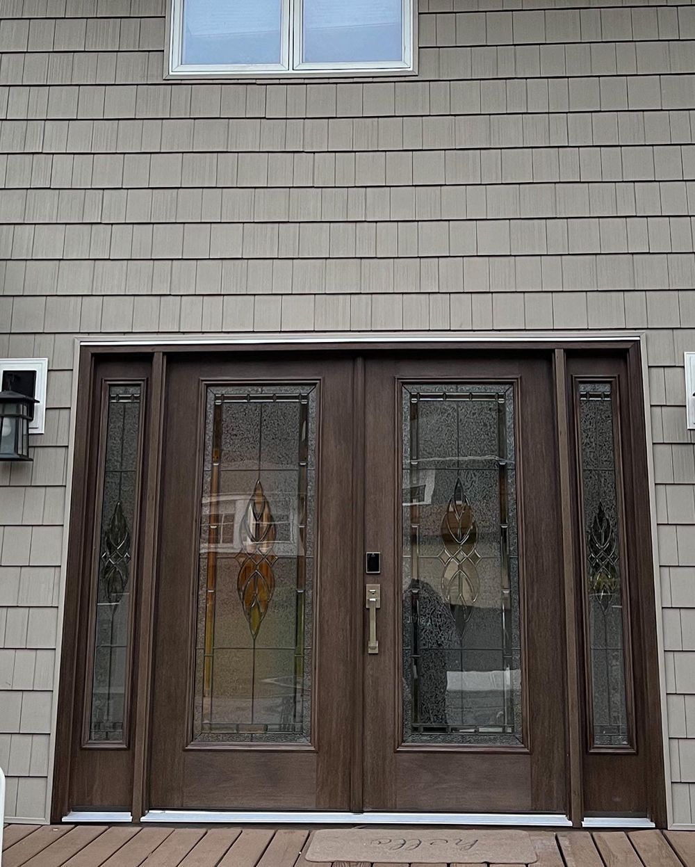 Brown double front doors with glass panes and side panels. Grey siding on house.