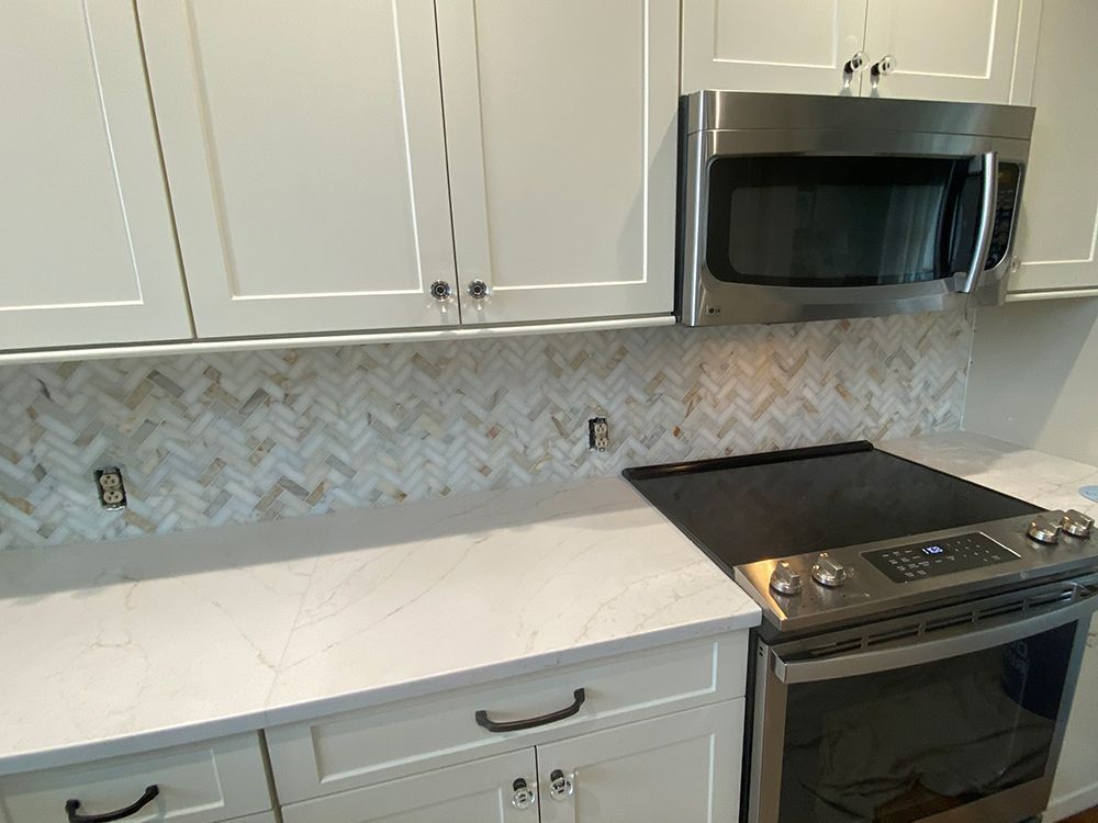 White kitchen cabinets with white countertop, and herringbone backsplash. Microwave and stove visible.