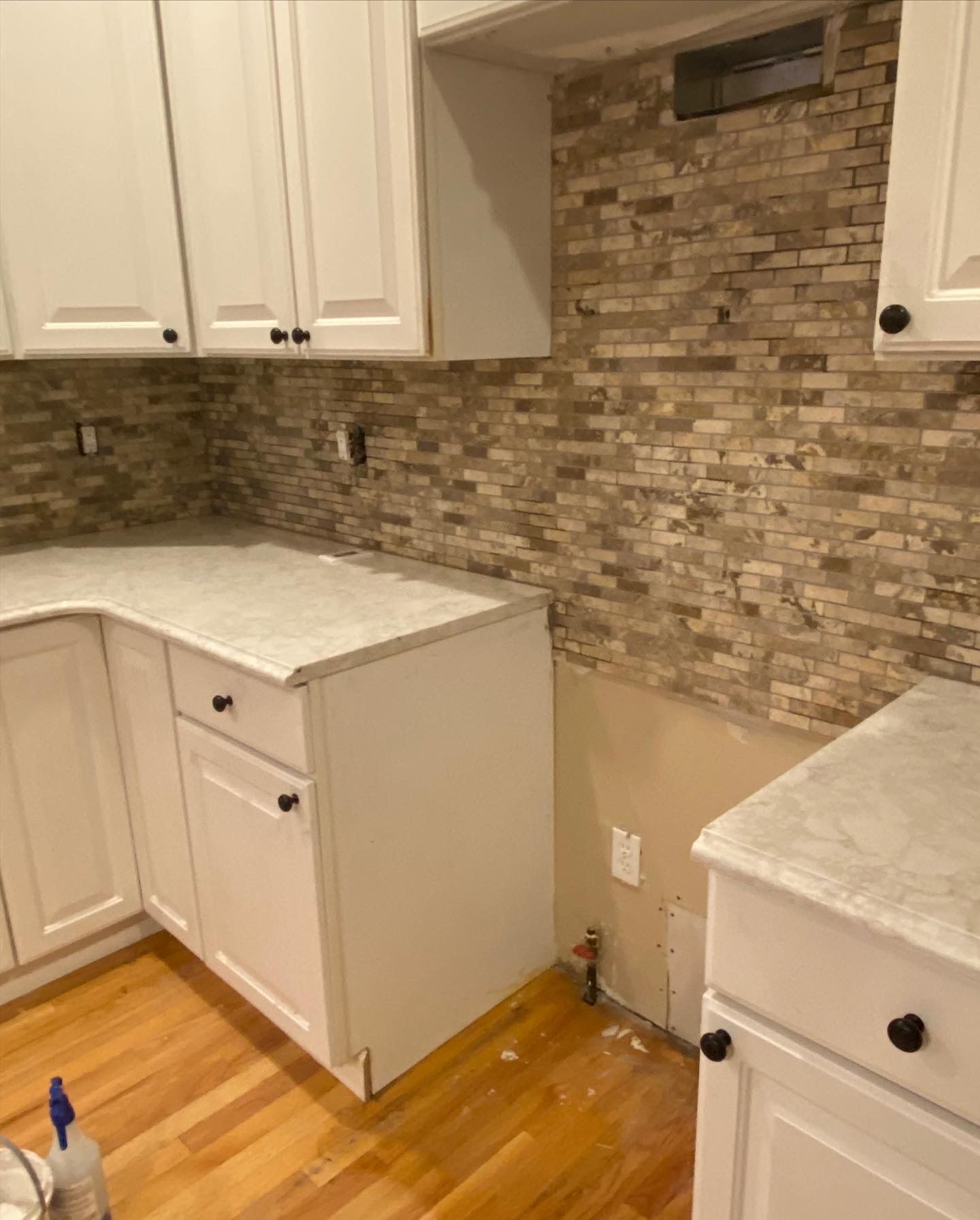 Kitchen undergoing renovation: white cabinets, countertops, and brick-style backsplash. Unfinished wall visible.
