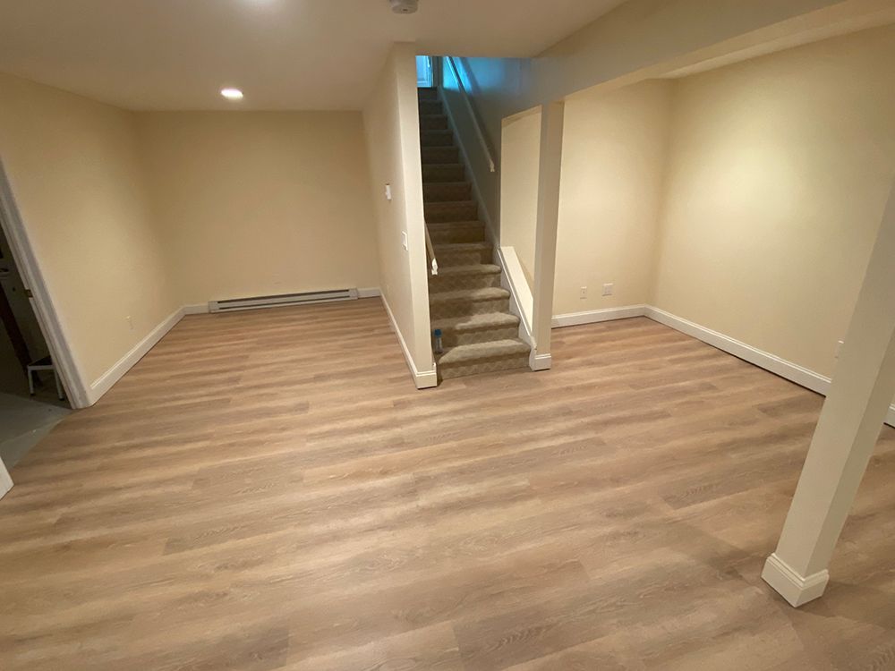 Empty basement room with light-colored flooring and walls. Staircase leads upwards.