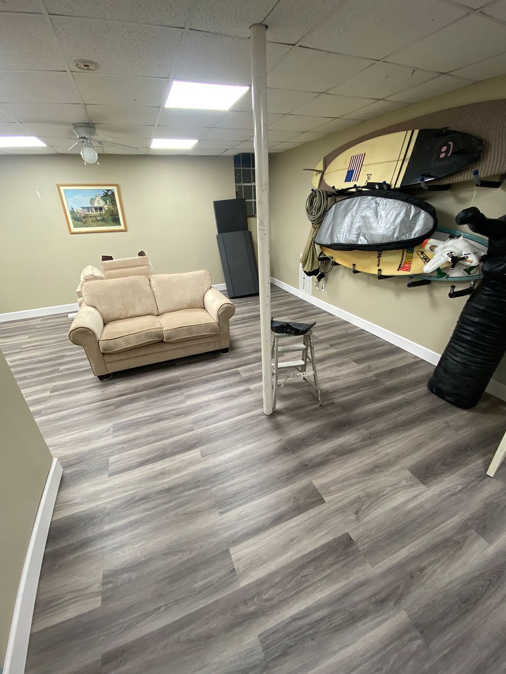 Basement with neutral wood-look flooring, beige walls, and a sofa. Surfboards hang on a wall.