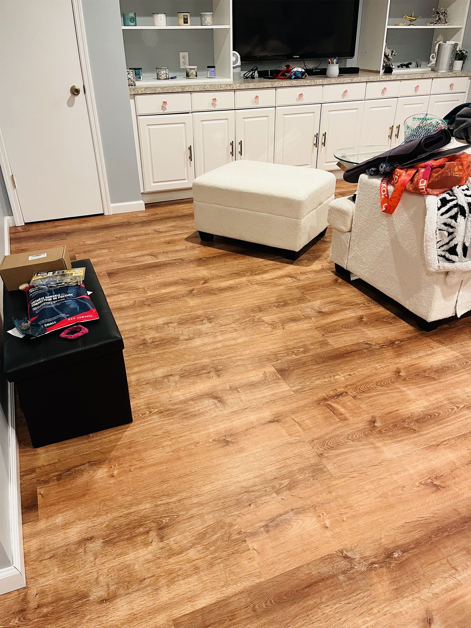 Wooden floor in a living room with white cabinets, ottoman, and black storage bench holding clothes.