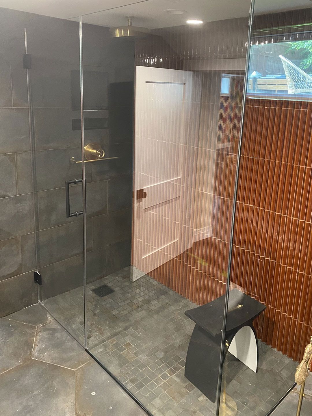 Modern bathroom with frameless glass shower and brown tiled walls. Door is off-white and a black bench is on the right.