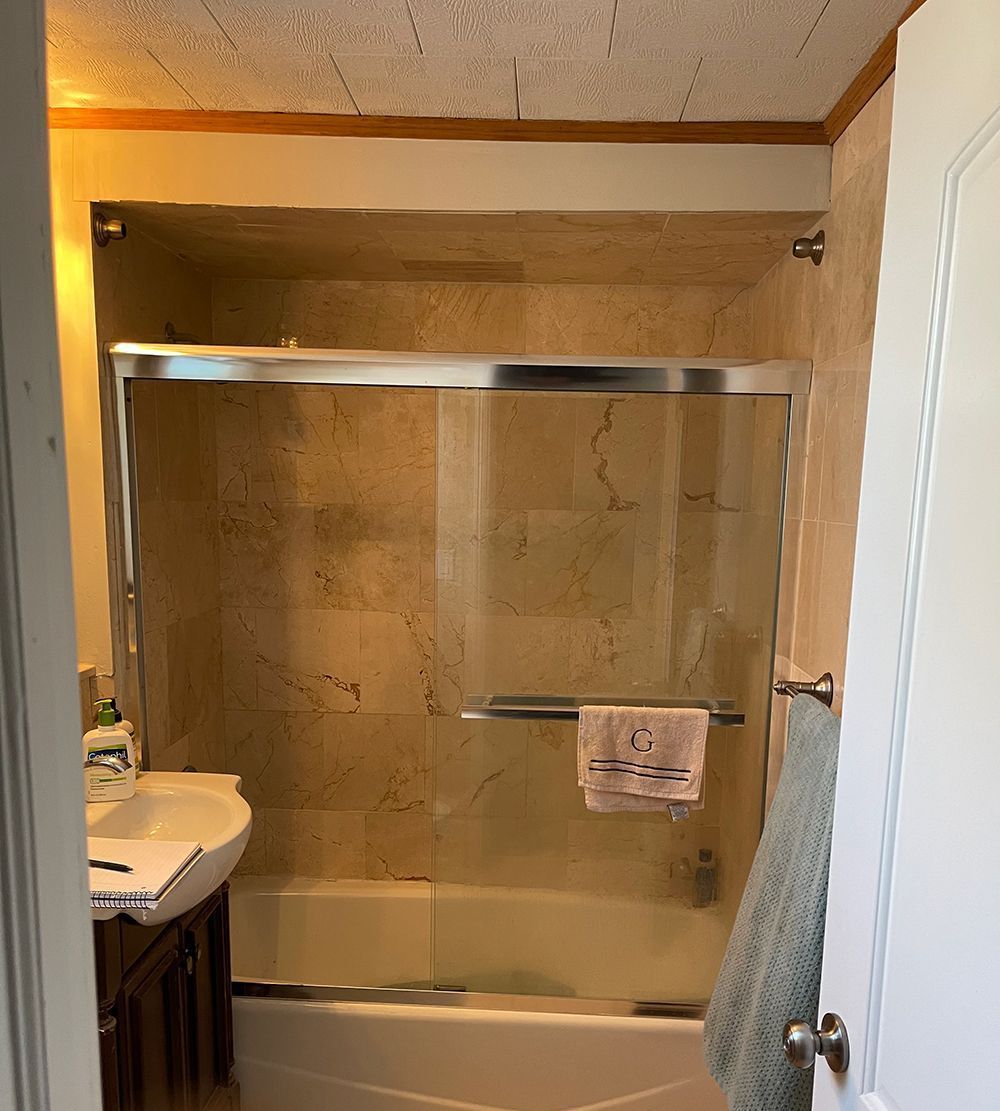 Bathroom with tan tiled walls, a glass shower door, and a white bathtub. A hand towel hangs on the door.