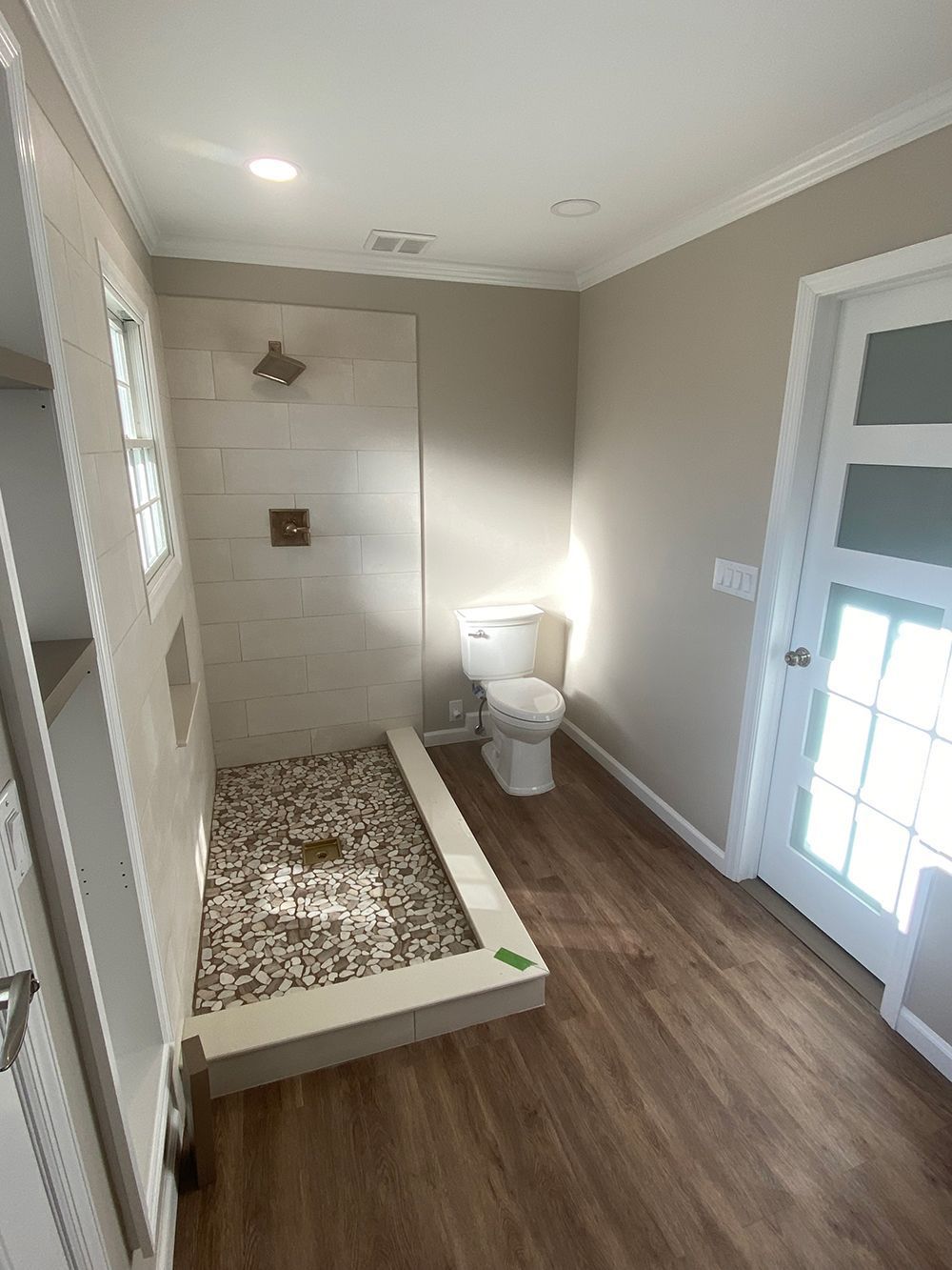 Modern bathroom with a stone-floored shower, toilet, and a partially glass door. Brown flooring and beige walls.