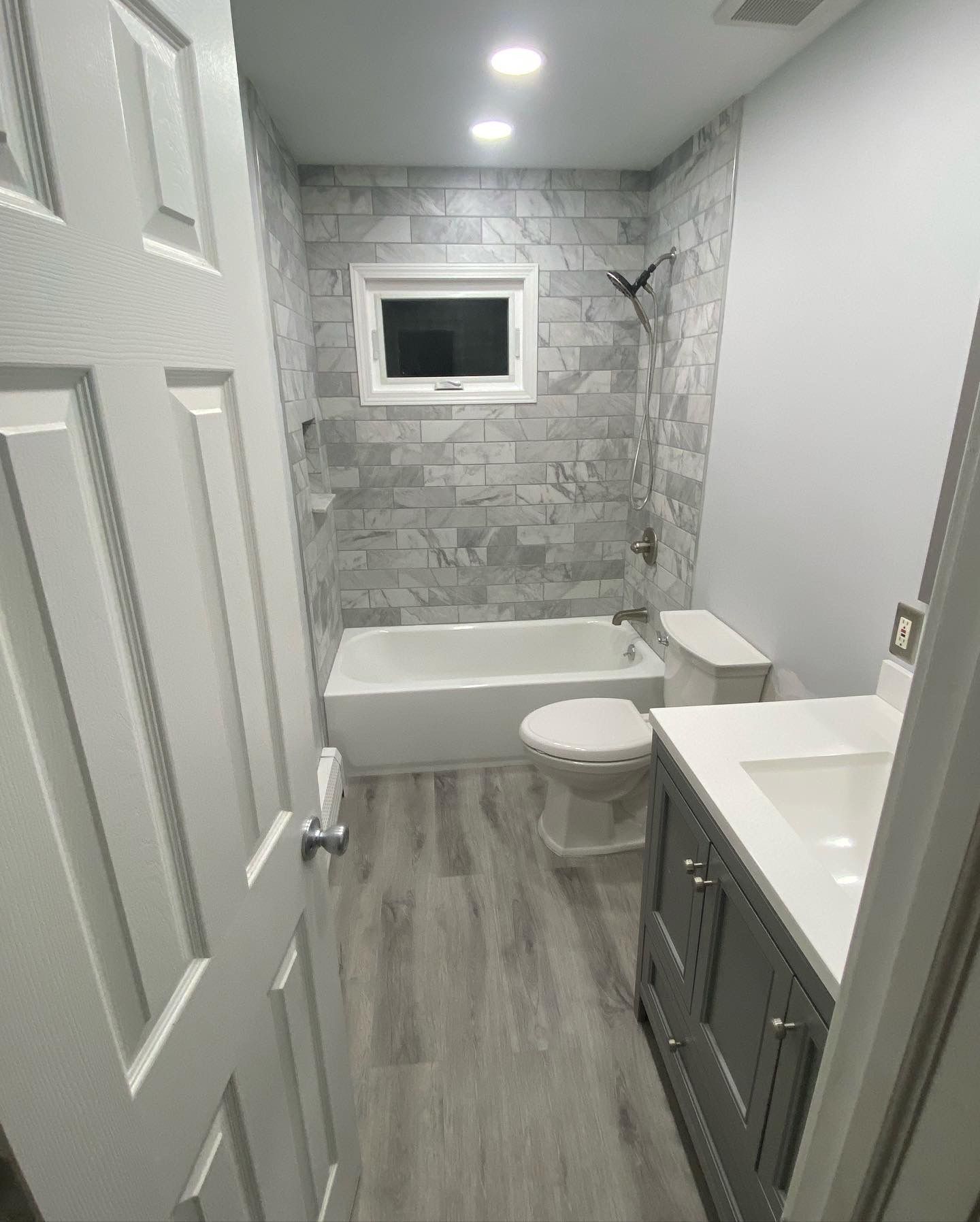 Bathroom with gray and white tile, white tub, toilet, and vanity, gray wood-look floor, and a small window.