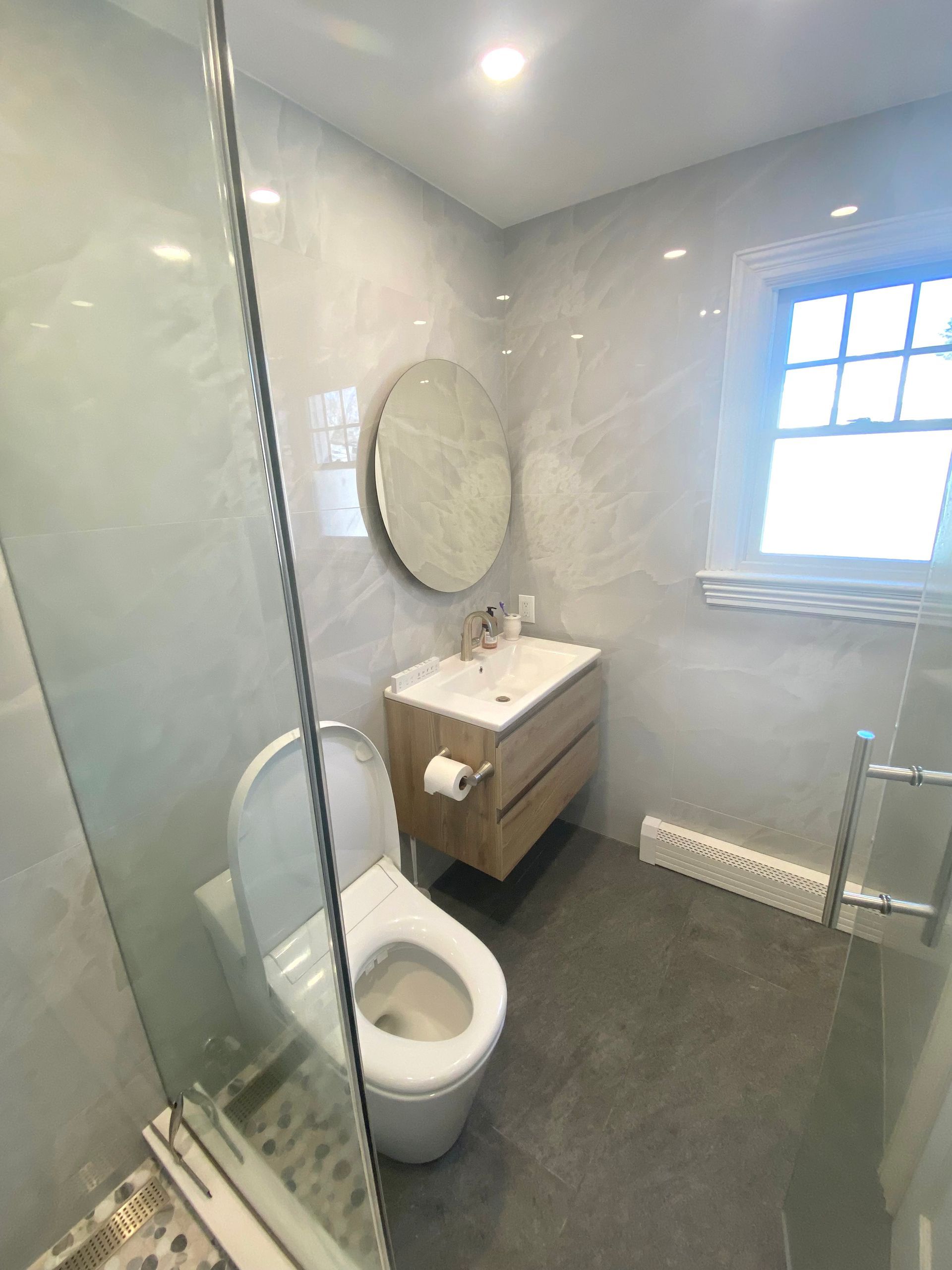 Modern bathroom with a toilet, sink, and round mirror. Gray and light wood tones.