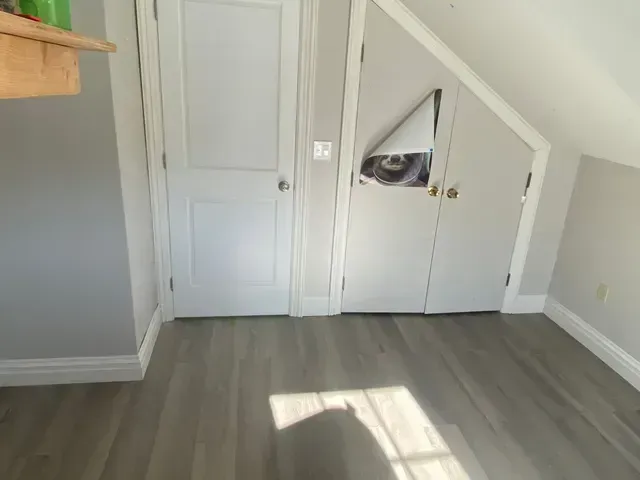 A room with gray walls and flooring, two white doors, one with a hole, and a slanted ceiling.