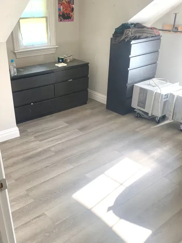 Bedroom with gray wood-look floor, black dressers, window, and air conditioner unit.