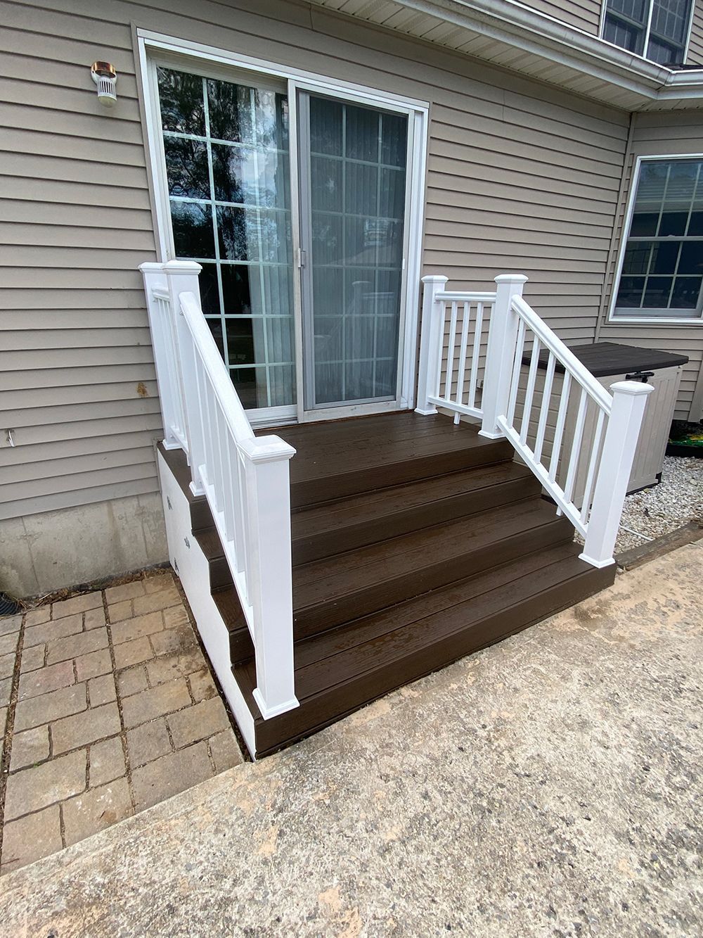 Brown steps with white railing leading to a sliding glass door. A brick patio is in the foreground.