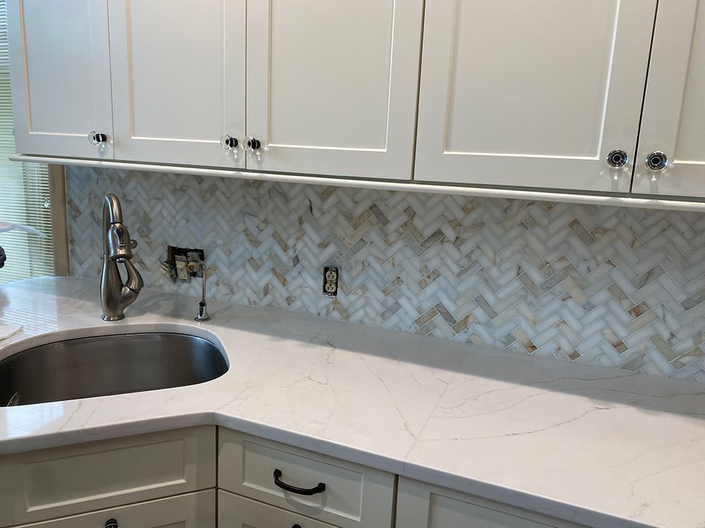White kitchen cabinets above a white countertop with a stainless steel sink and herringbone tile backsplash.