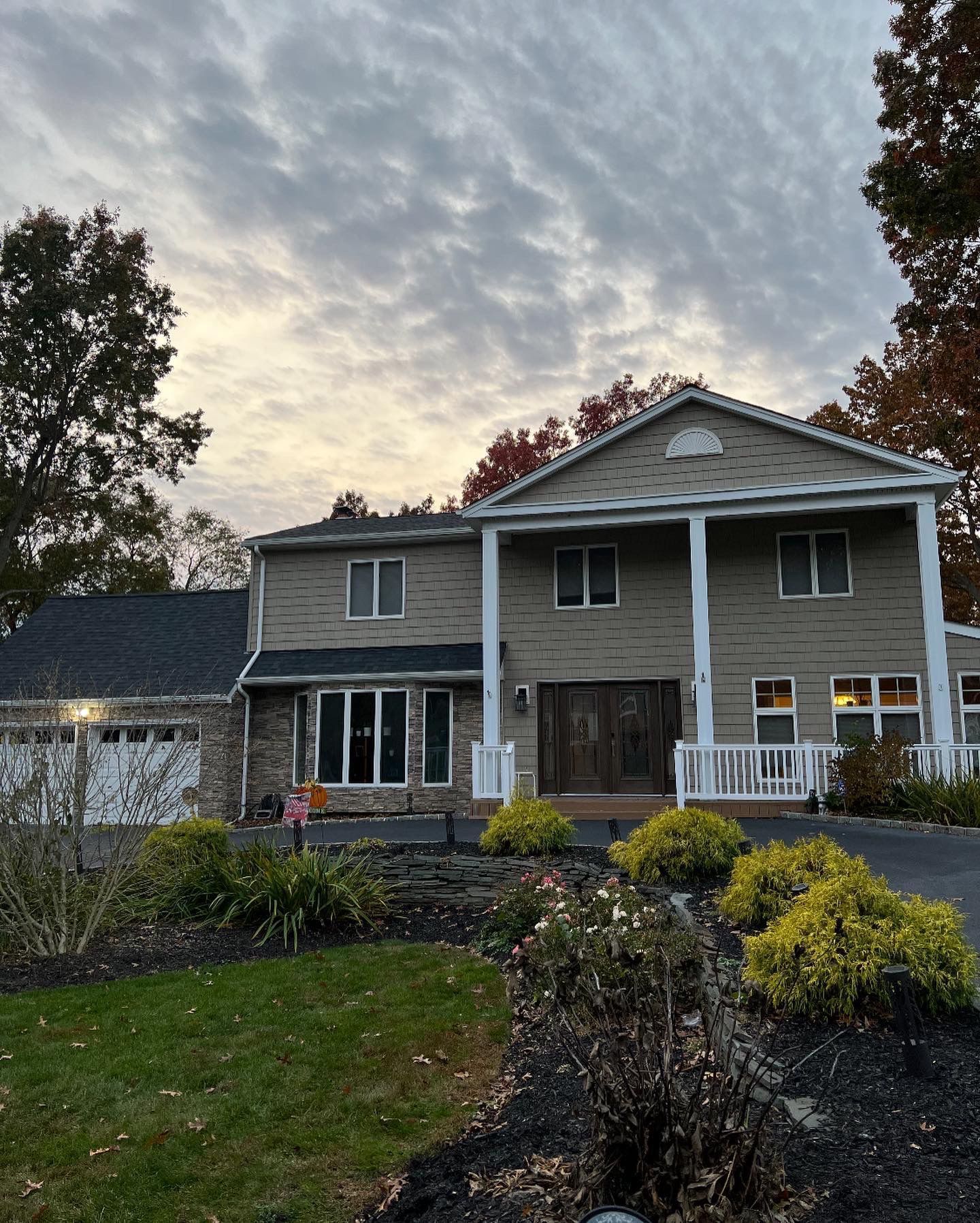 Two-story house with columns, a dark roof, and cloudy sky. Landscaping in front includes shrubs and a driveway.