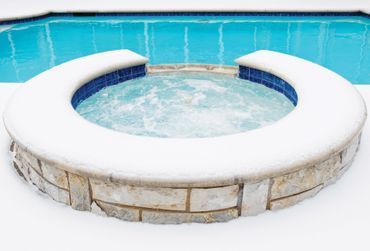 A hot tub surrounded by snow next to a swimming pool.