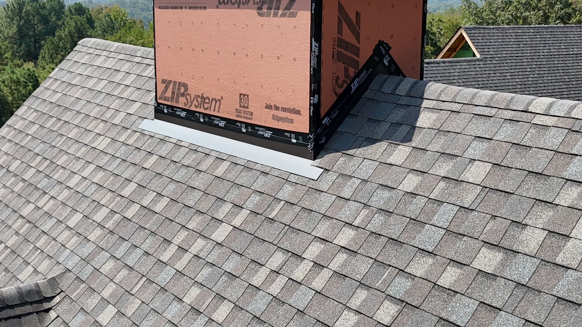 Chimney on a roof with gray shingles and metal flashing.