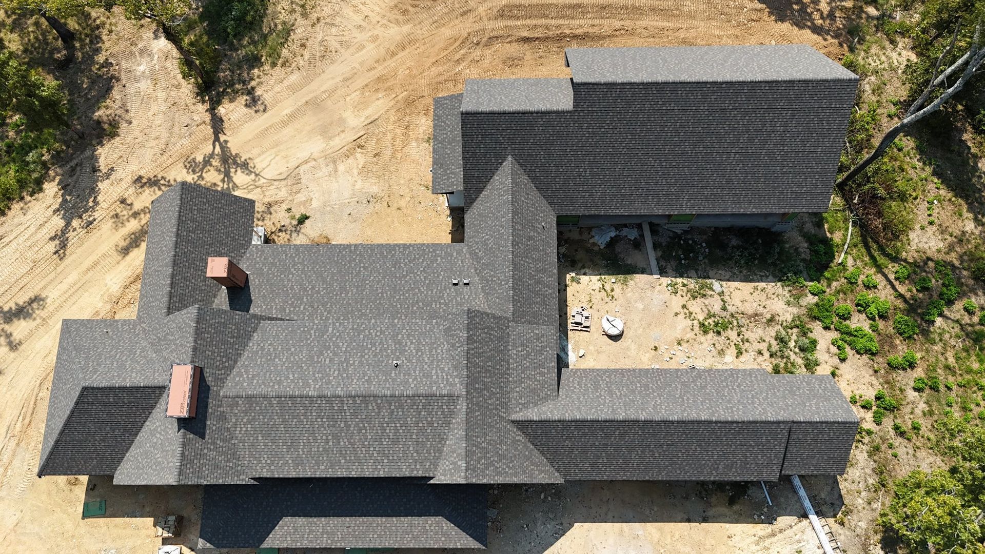Aerial view of a dark gray shingled roof under construction. Two red brick chimneys, on dirt ground.