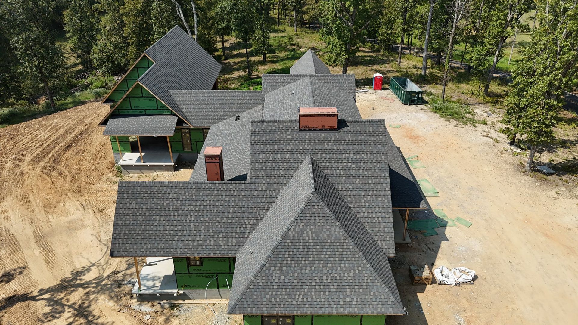 Aerial view of a house under construction with a gray shingle roof in a wooded area.
