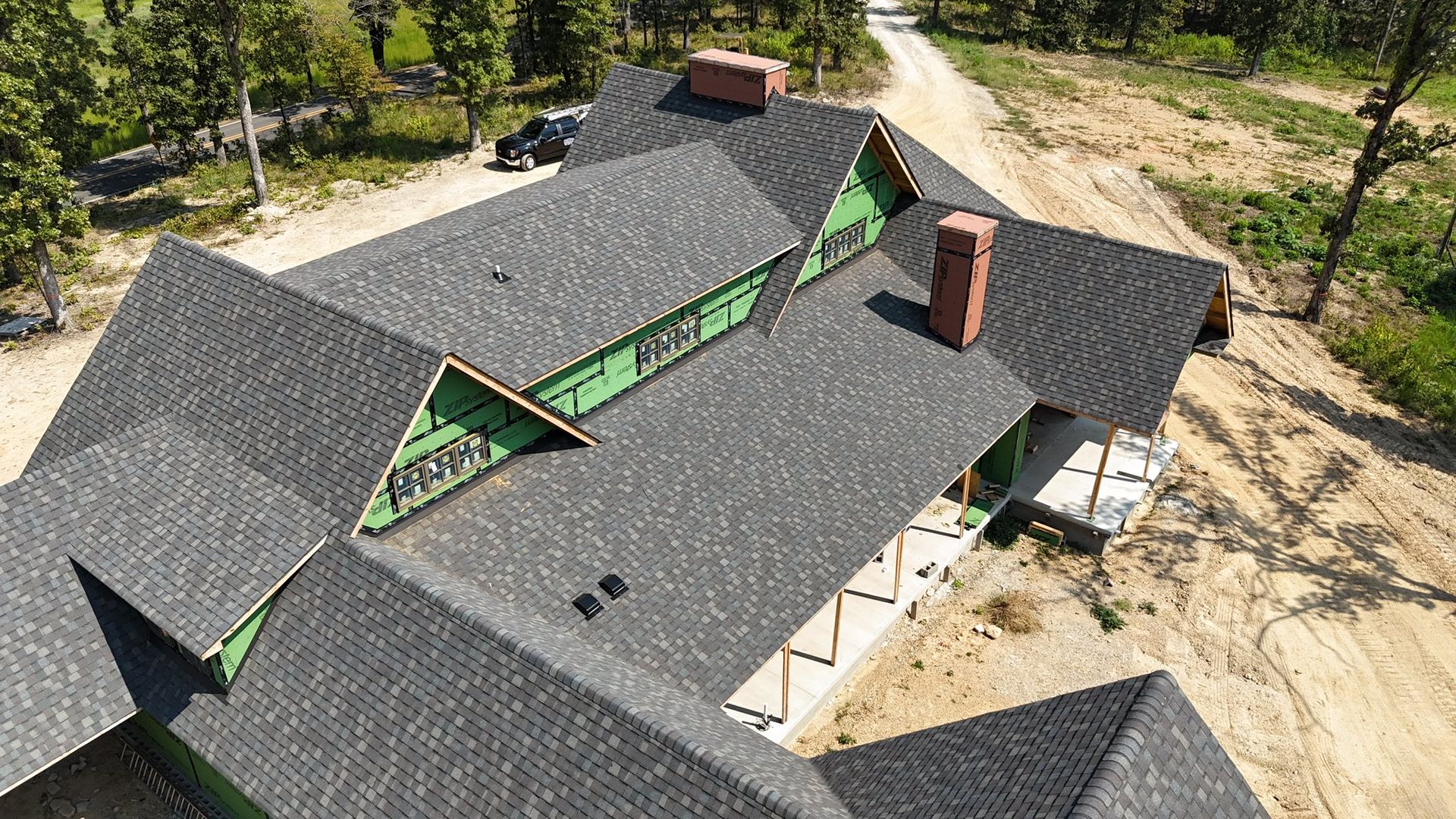 Dark gray shingled roof of a house under construction; brick chimneys, green framing, and dirt ground visible.