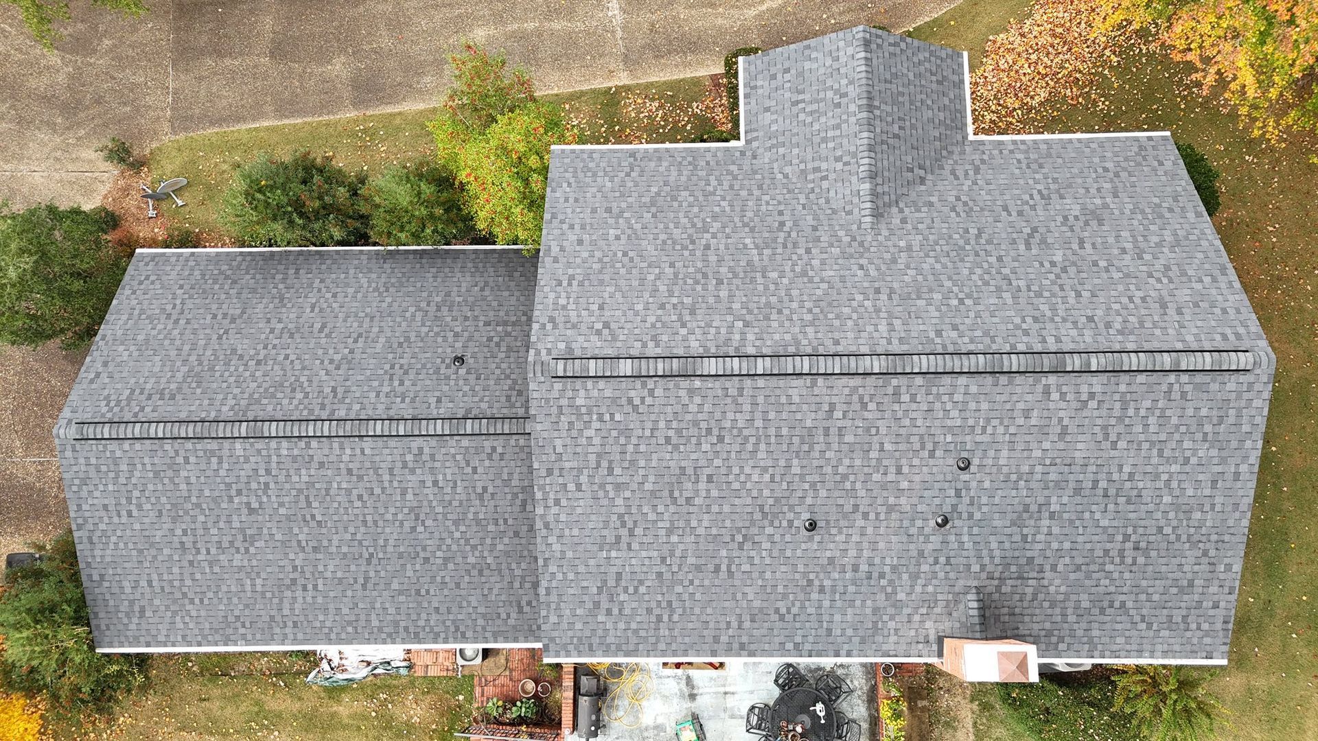 Aerial view of a gray asphalt shingle roof on a house with a chimney, surrounded by trees and grass.