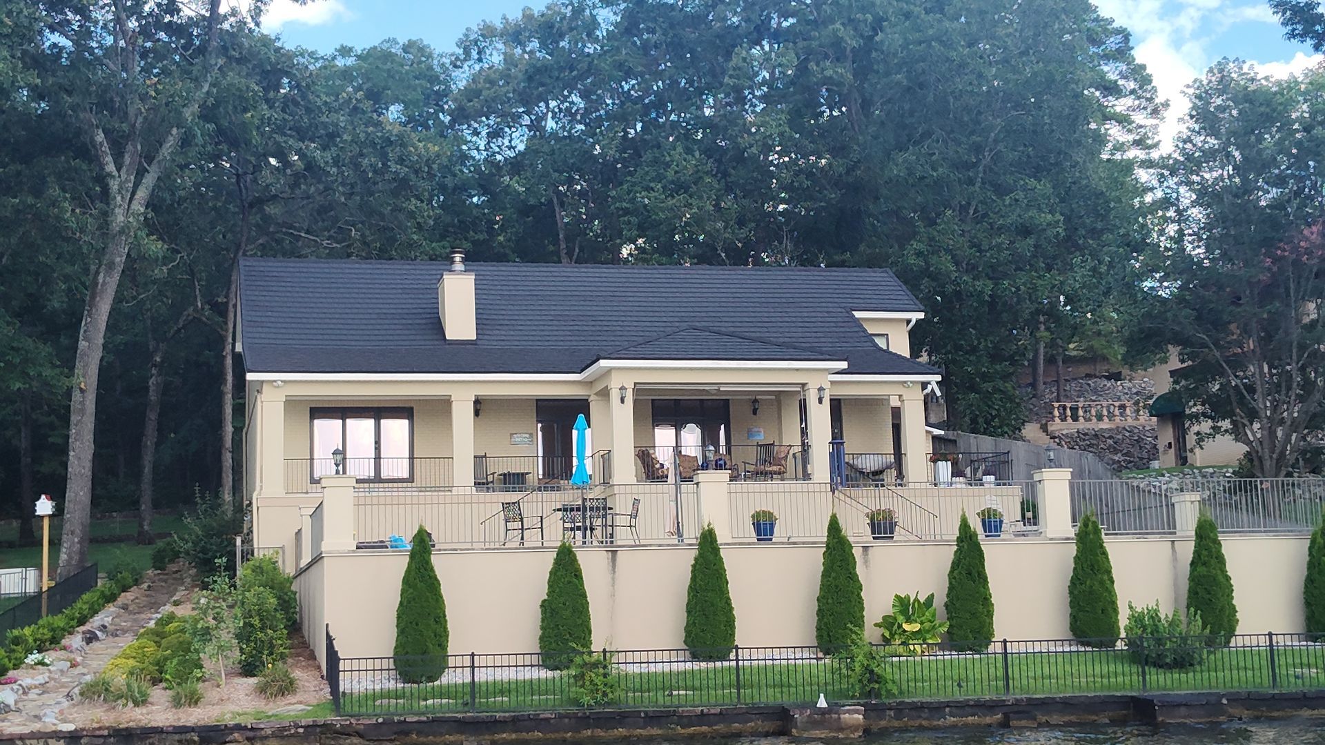 Beige house with black roof, outdoor seating, and small trees in front, set against a backdrop of trees.