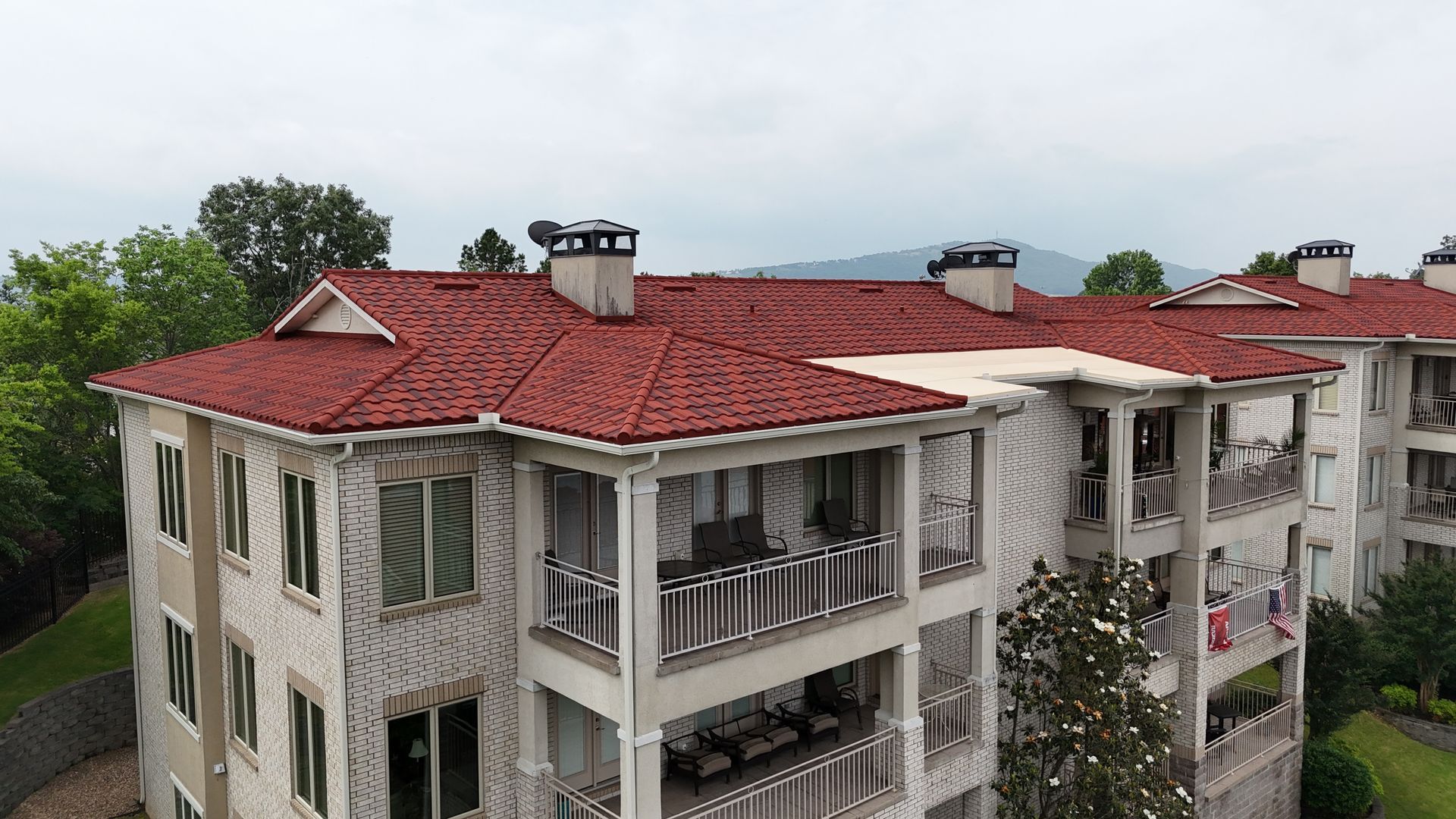 Apartment building with red tile roof under a cloudy sky; green trees and mountains in background.