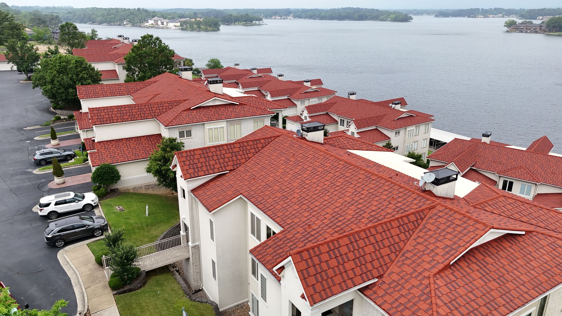 Row of white buildings with orange tile roofs on a lake, cars parked on the side.