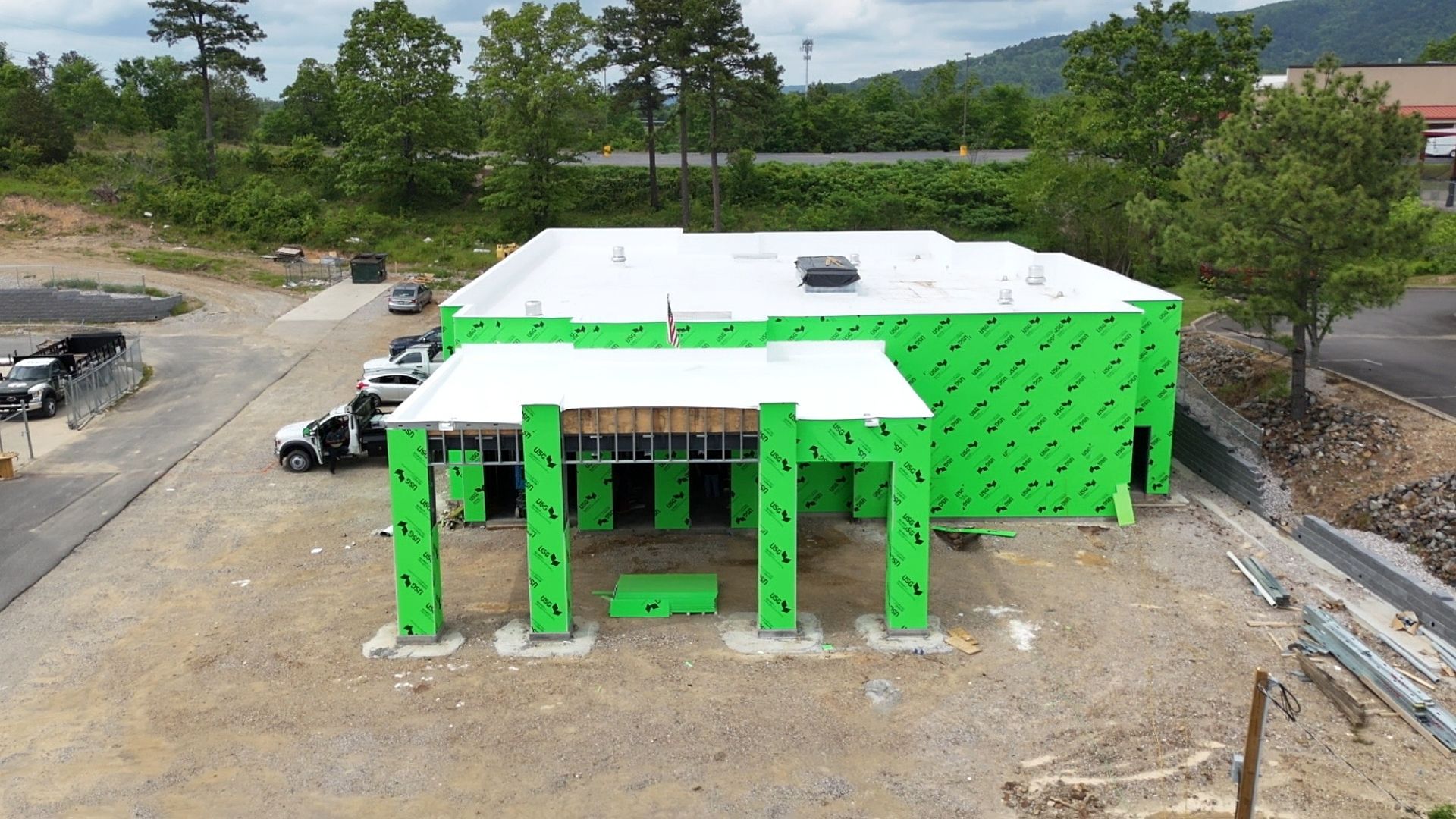 Building under construction, green wrap, white roof, outdoor setting, trees and vehicles.