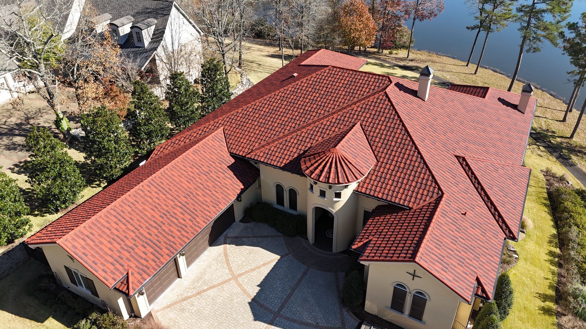 Aerial view of a large beige house with a red tile roof, overlooking a lake and trees.