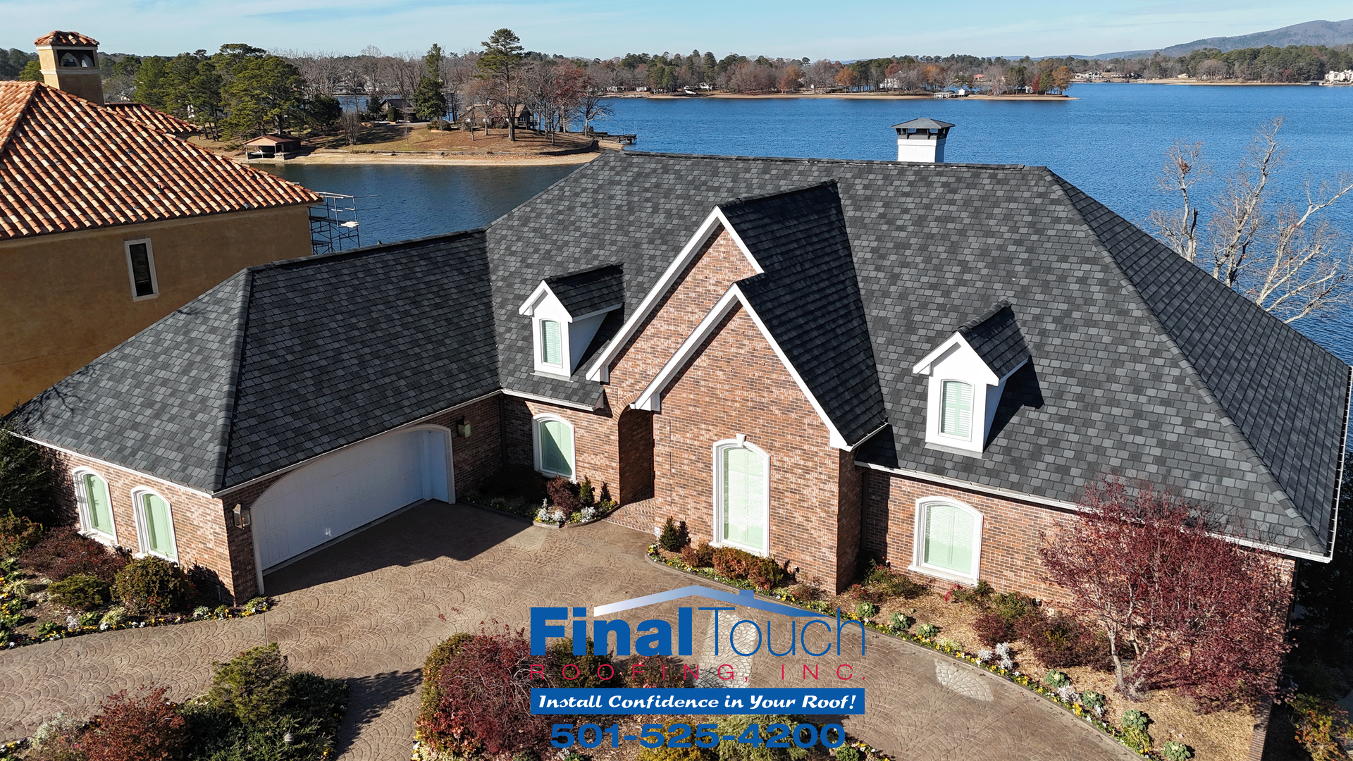 A brick house with a dark shingle roof sits near a lake on a sunny day.